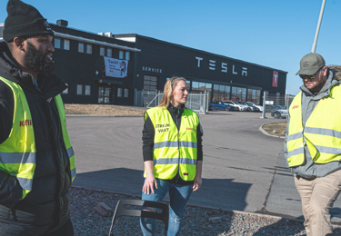 Tre fackliga medlemmar i reflexvästar som står utanför en byggnad med Teslas logotyp på
