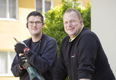 Två elektriker i en somrig miljö som tittar in i kameran och ler