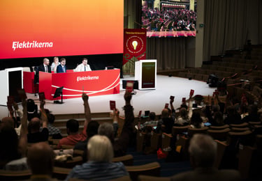 Elektrikernas kongress med uppsträckta händer som håller i röstkort i förgrunden och med de tre personerna i presidiet i bakgrunden